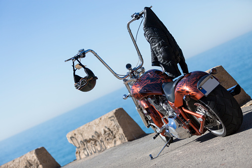 customized chopper motorcycle with a helmet and a leather black jacket hanging on the handlebar over a sea and sky background at sunrise - focus on the helmet