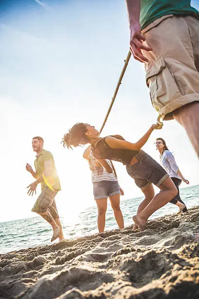Group of multi ethnic friends, teenagers, playing together on the beach at dusk. They are dancing and playing to limbo during a summer vacation. The sunlight on the background is warm and the group of friends is enthusiast about the game