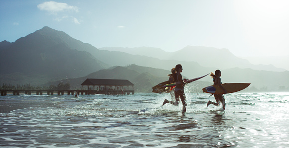 Three people on a beach, running into the ocean with surfboards in a tropical climate, with mountains in the background.