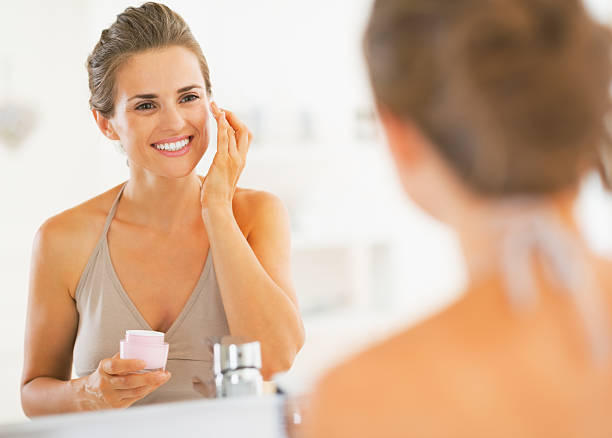 felice giovane donna applicando crema in bagno - moisten foto e immagini stock