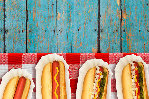 Four hotdogs on red and white gingham napkin from plain to all the fixin's. Condiments include ketchup, mustard, relish and onions. All hotdogs served in sanitary white paper fluted hot dog trays. The blue weathered picnic table provides copy space in a summer holiday red, white and blue theme for July Fourth, Memorial Day and Labor Day picnics.