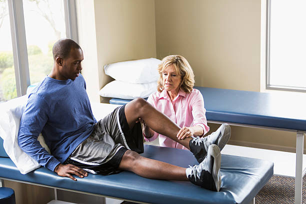 Physical therapist examining patient A female physical therapist examining a young African American man on a treatment table.  He is sitting on the table with his legs stretched out in front of him.  The physical therapist, a senior woman, is examining his knee.  They are both looking at his bent knee with serious expressions on their faces. orthopedist stock pictures, royalty-free photos & images