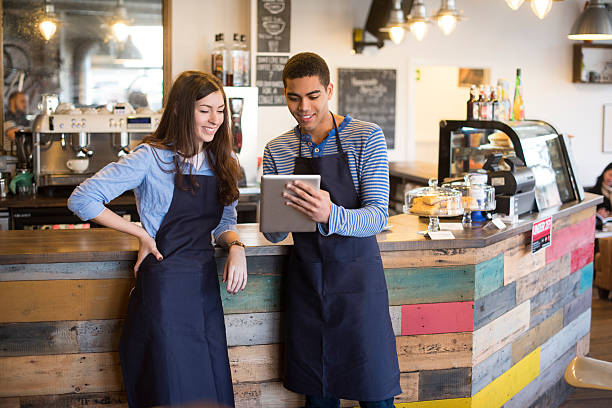 coffee shop workers using a digital tablet A young coffee shop waiter and waitress are standing in front of the counter  and using technology  . The young man holds a digital tablet and is updating the cafe's social media page . They are both wearing  blue aprons . first job photos stock pictures, royalty-free photos & images