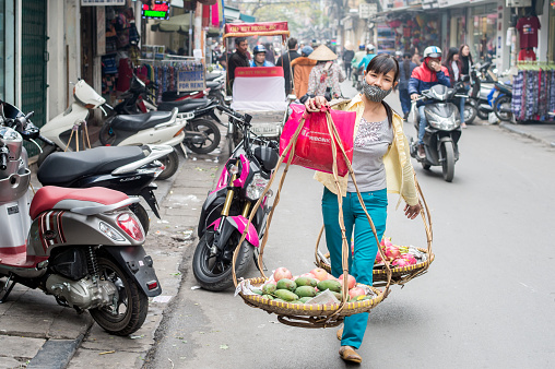 Hanoi, Vietnam – February 10, 2015: Vietnamese woman selling fruits from two baskets hanging on a carrying pole in the old quarter of Hanoi. The 36 old streets and guilds of the old quarter are a major tourist attraction.