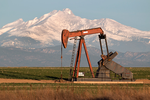 In the morning, an oil derrick pumps in the in green agricultural fields in the Niobrara Oil Shale Formation off Interstate 25. The area is located in Weld County north of Denver and just east of the town of Erie in the Wattenburg field with the snow covered Mount Meeker and Longs Peak, part of the snowy Rocky Mountains in the distance. 