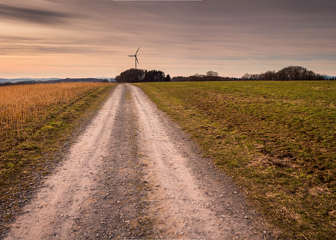 Spring landscape with a path and a wind turbine in Germany. The Path in the foreground leads to the windturbine in the background. The turbine is located behind a small forrest. On the right hand side there is a field.