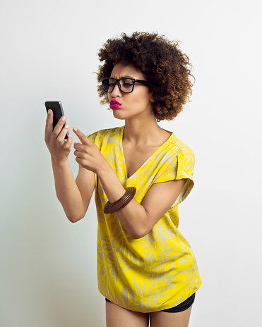 Portrait of irritated afro american young woman wearing yellow top, shorts and nerd glasses, using a smart phone. Studio shot, white background.