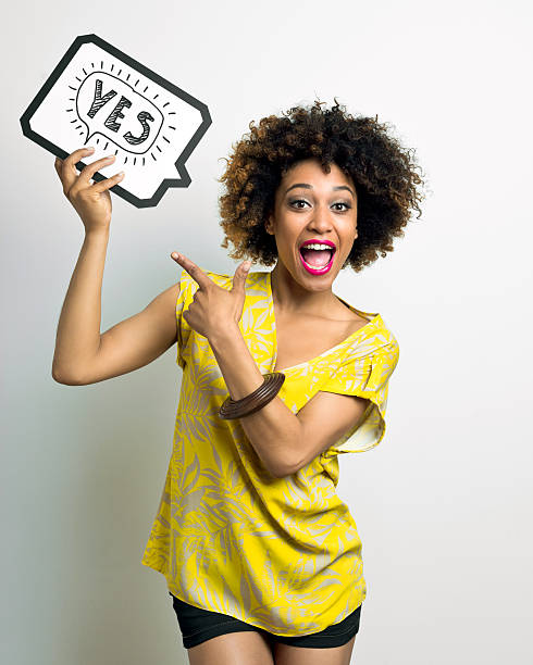 Excited Afro American Woman with speech bubble Portrait of excited afro american young woman wearing yellow top and black shorts, holding thought bubble with drawn word yes above her head, pointing with index finger and shouting at camera. Studio shot, white background. yes single word stock pictures, royalty-free photos & images