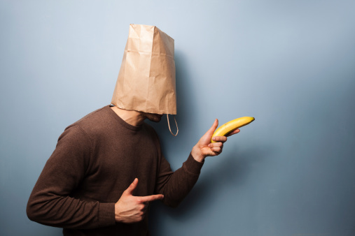 Young man with a paper bag over his head is using a banana as gun