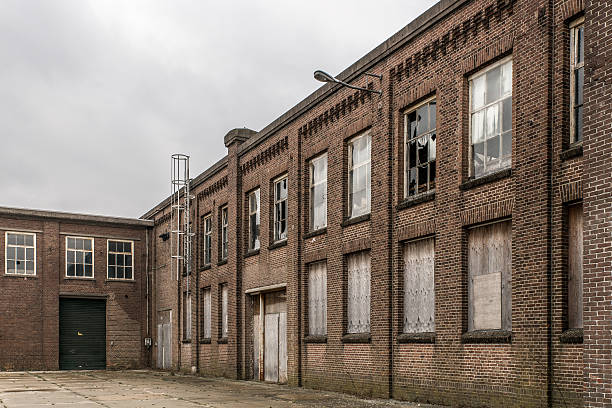 abandoned factory Wide angle view of a brick facade of an old and abandoned factory with broken glass windows and some windows covered witch wooden plates. In front of the building concrete slabs lay on the ground detroit ruins stock pictures, royalty-free photos & images