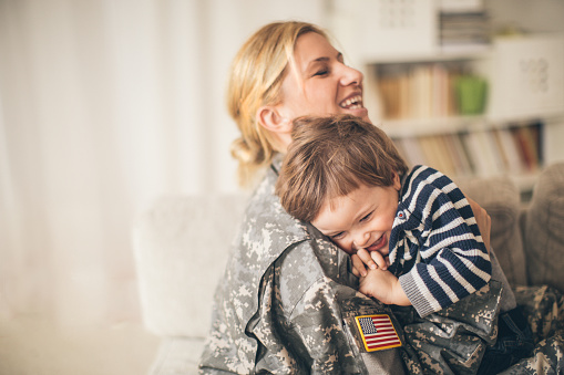 Photo of smiling young woman soldier and her son seeing after a long time and enjoying in their living room. Woman is sitting on the sofa and hugging him