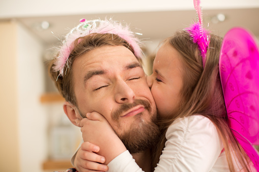 Father and his cute daughter in kitchen taking selfies with smartphone. Personal perspective. Little girl wearing fairy costume and tiara, embracing and kissing her dad.