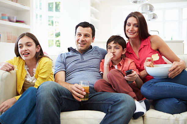 Hispanic Family Sitting On Sofa Watching TV Together Hispanic Family Sitting On Sofa Together Watching TV After Work And School family eating potato chips stock pictures, royalty-free photos & images