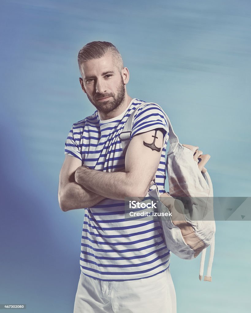 Bearded sailor man wearing striped t-shirt Portrait of confident bearded sailor man with anchor tatoo on shoulder wearing white and blue striped clothing, holding backpack. Standing with arms crossed against blue background, looking at camera. Studio shot, one person. Men Stock Photo