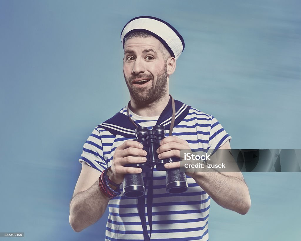 Bearded man in sailor style outfit holding binoculars Portrait of surprised bearded sailor man wearing white and blue striped clothing and sailor hat, holding binoculars in hands. Standing against blue background. Studio shot, one person. Sailor Stock Photo