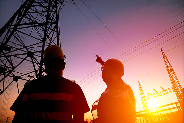 A low-angle shot captures two workers scaling a towering red electrical transmission tower against a pale, overcast sky. The tower's intricate steel lattice structure forms a stark geometric pattern, contrasting with the soft, diffused light. The workers, clad in safety harnesses and work gear, appear as small figures against the massive framework, emphasizing the scale of the structure and the height at which they are working. Thin electrical wires stretch horizontally across the frame, adding to the industrial aesthetic. The image highlights the challenging and potentially dangerous work of maintaining essential infrastructure.
