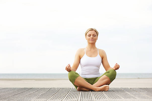 Beautiful young woman sitting in yoga lotus pose at beach Portrait of a beautiful young woman sitting in yoga lotus pose at the beach yoga lotus position meditating women stock pictures, royalty-free photos & images