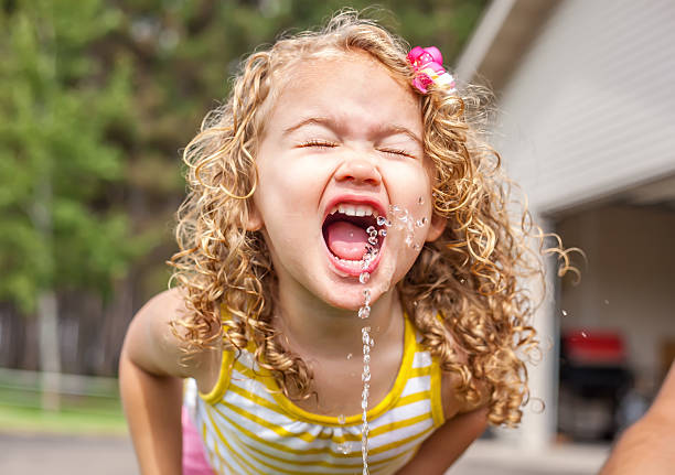 Curly Haired Little Girl Drinking Water From Hose Fountain Little girl drinking from a fountain made by the garden hose, on a hot summer morning. She has curly blonde hair and is wearing a yellow and white striped tank top. The adult's arm who is holding the hose for her is visible in the bottom right corner. preschooler caucasian one person part of stock pictures, royalty-free photos & images