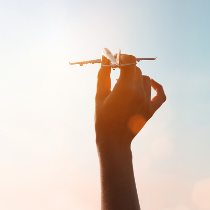 Silhouette of a woman hand holding a metal airplane model ready to take off and fly toward a pastel colored daylight sky with shining sunlight and lens flare. Freedom, travels and dreamlike lifestyle.