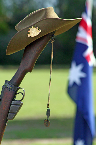 Portrait shot (with room for copy) of an upturned, vintage Australian Army 303 rifle, a soldier's dogtags, slouch hat with the famous rising sun badge and an Australian Flag flying in the background during an ANZAC Day memorial service.