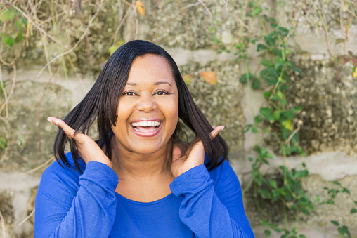 Portrait of a confident, mature African American woman with a big smile, and hands in her straight hair.  She is standing outdoors in front of a stone wall, wearing a royal blue shirt.