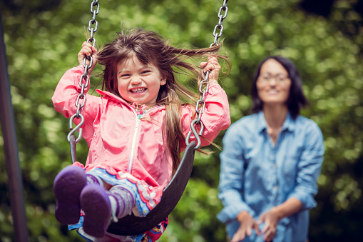 Little girl laughing in mid swing as her mom stands in the background ready to push her again.