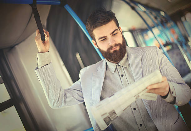 Early 30's handsome businessman reading newspapers in public transport on his way to work. He's dressed in light colored suit and has full grown beard.