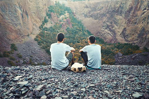 Two tourist young men sitting on rocky cliff and enjoying beautiful view