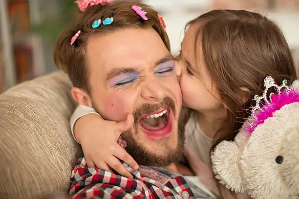 Cute daughter and her father having princess time. Father looks funny, wearing makeup and tiara, holding bear toy wearing tiara too. Girl embracing him and kissing. Bonding time.