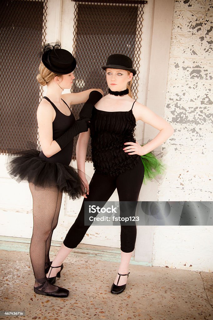 Pair of jazz ballet dancers wearing costume pose for camera. Two Caucasian attactive young teenage ballet jazz dancers wearing black costumes with hats.  They pose outside an abandonded store front in a ghost town.  Both have blonde hair and wear felt hats.  One wears a net tutu, stockings and ballet shoes.  One wears black leggings with tap shoes.  She has a green tuff of netting on her hip.  And wears a black chocker.   Canon DSLR. 2015 Stock Photo