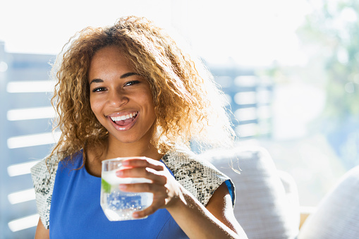 An attractive young African American woman sitting by a bright sunny window, drinking a glass of water with a slice of lime in it.  She is laughing with her mouth open.