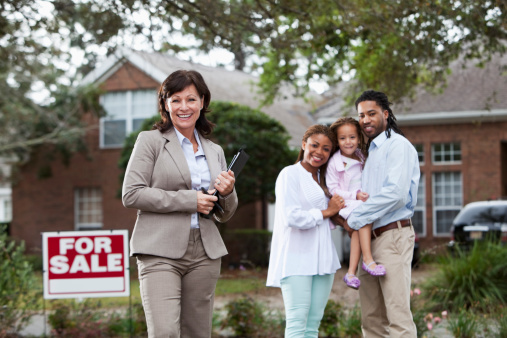 Real estate agent (50s) with young family outside house with For Sale sign.  Main focus on agent.