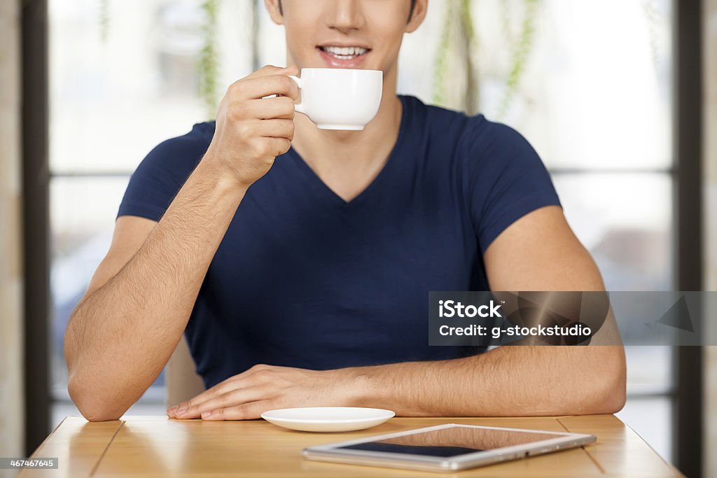 Drinking coffee at restaurant. Cropped image of cheerful young man drinking coffee at the restaurant and smiling while digital tablet laying on the table Adult Stock Photo