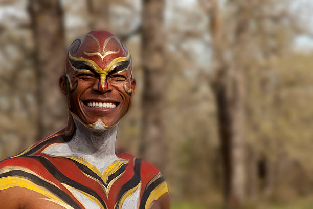 African hunter or warrior in full body paint smiling outdoors in a forest area. Portrait style. Red, black, yellow, and white paint on his torso and face.  He is looking at the camera.  Many trees in background.