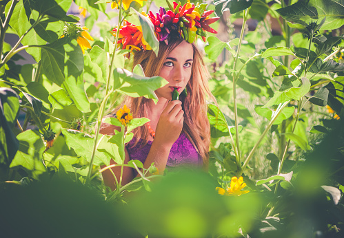 Long haired, blonde teen girl wearing a wreath of fresh peppers on her head, while in the garden. She's holding a green jalepeno pepper up to her lips, looking up and away from the camera. She's surrounded with vegetation of green leaves and yellow sunflowers. It's a sunny summer day. Horizontal composition.