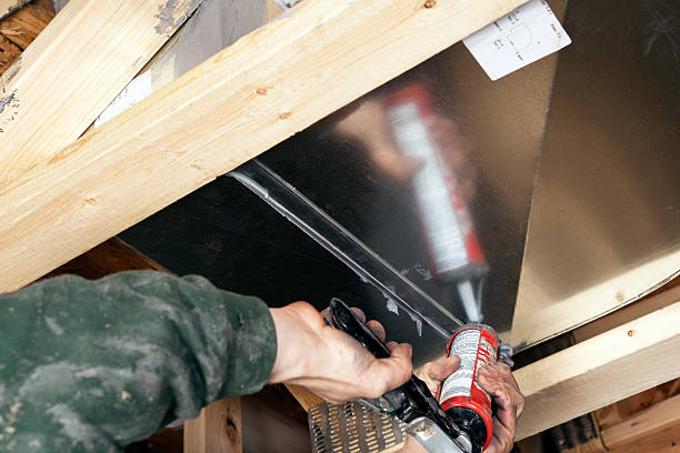 Dirty male hands are using a caulk gun to apply sealant to a house air duct. Caulking ducts prevents air-conditioned or heated air from escaping the duct and prevents ambient air from entering the system. This is a new practice in homebuilding to increase energy efficiency, eventually it may become standard or mandatory.