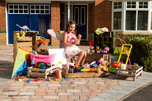 A happy little girl sits infront of her house with some old toys to sell. She is smiling at the camera as she plays with one of them. Her house can be seen in the background.