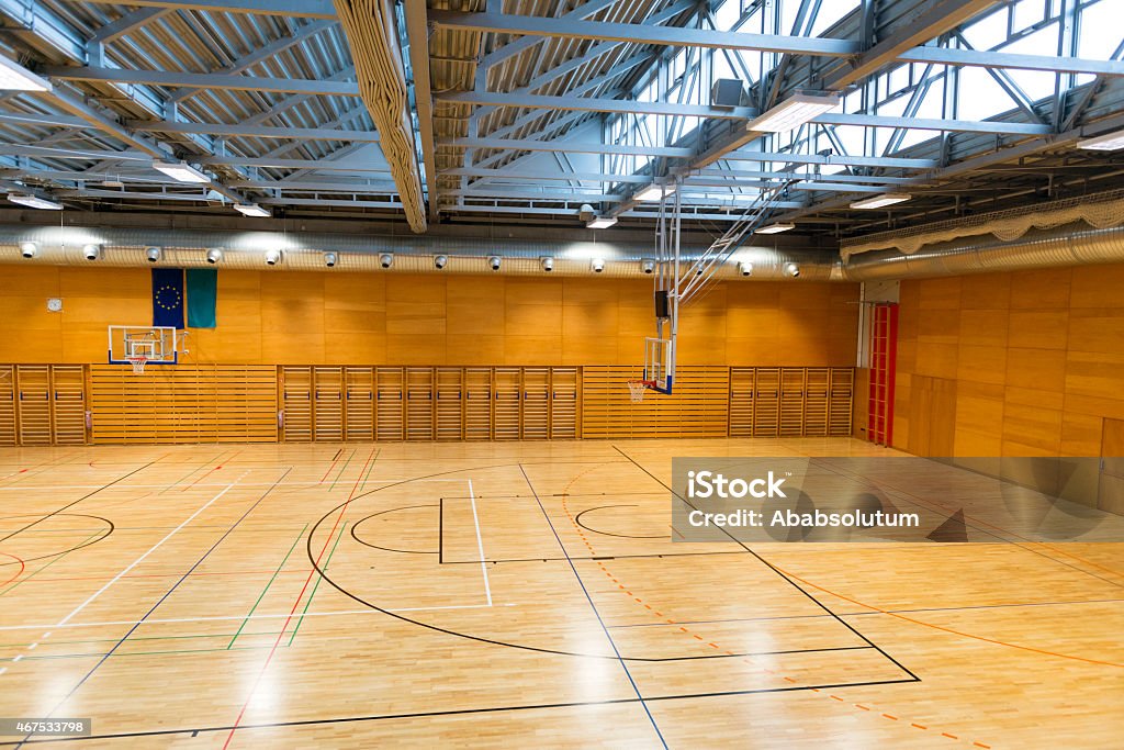 Empty Sports Hall, Metal Roof, Wall Bars, New Parquet, Europe School gymnasium with metal roof, basketball backboards, wall bars, European flag on the wall, daylight from roof windows. Wooden walls and beautiful new parquet with lines and curves for different sports(basketball, volleyball, football). Elevated view. Brown and gray colors dominate. Nikon D800, full frame, XXXL. School Gymnasium Stock Photo