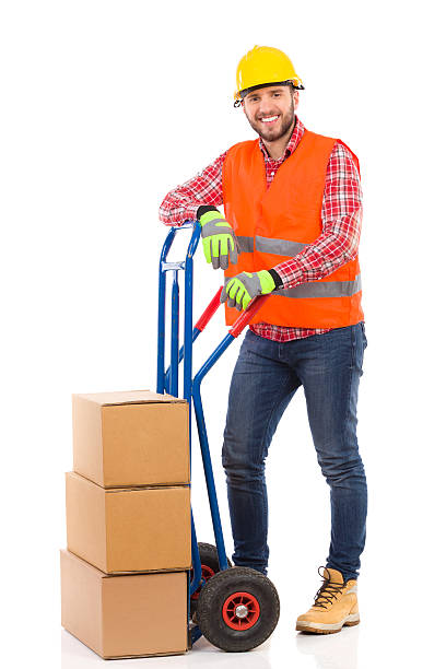 Smiling mover posing Happy man in yellow hardhat and orange reflective vest posing with a delivery cart and looking at camera. Full length studio shot isolated on white. mover delivery person box hand truck stock pictures, royalty-free photos & images