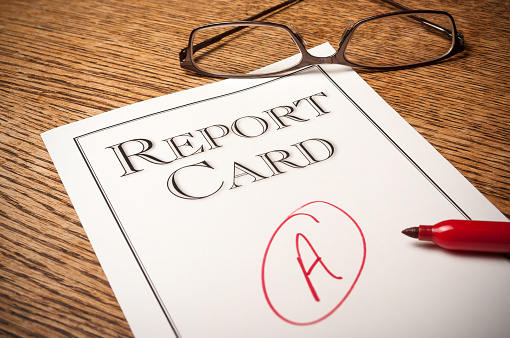 Report card on an oak desk with a red “A” written on it . A red pen in the foreground and glasses in the background.