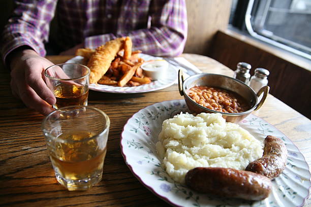 British pub food POV of table with man, two drams of whiskey and two plates of traditional British food: bangers and mash and fish and chips. pub food stock pictures, royalty-free photos & images