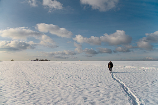 Footprints of a man with Jeans and a leather jacket walking pensively on a big bright white snowy field. Far away on the horizon there are some trees under an atmospherical heaven.