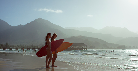 Two women standing side by side at the waters edge in a tropical climate carrying surfboards, looking out into the ocean with a pier and mountains in the background.