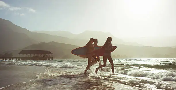 Three people on a beach, running into the ocean with surfboards in a tropical climate, with mountains in the background.