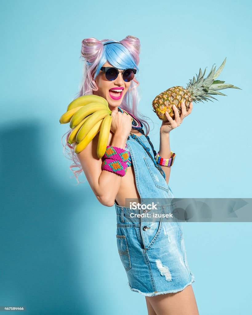 Pink hair girl in manga outfit holding pinapple and babanas Portrait of cheerful manga style pink hair young woman wearing denim coveralls and sunglasses. Standing against blue background, holding pineapple and bananas in hands and laughing at camera. Studio shot, one person. Bizarre Stock Photo