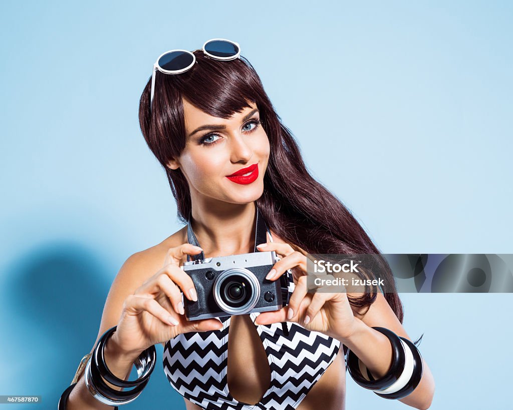 Smiling young woman wearing swimsuit taking picture using camera Summer portrait of glamour brown long hair young woman wearing black and white swimsuit taking picture using camera, smiling at the camera. Standing against blue background. Studio shot, one person. 2015 Stock Photo