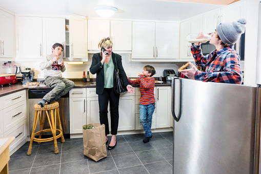A business woman and mother returns home with groceries to find her kids raiding the fridge and cupboards for food  She is on the phone with a client while her sons tugs at her sleeve.  A typical scene for women balancing business and family life.