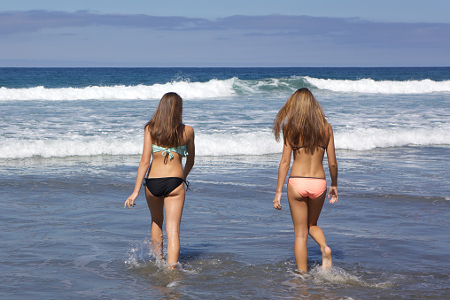 Two Teenage Girls in Bikinis going into the water at the beach. They are photographed from the back.  Both girls have long brown hair.    It is a beautiful summer day in San Diego, California 