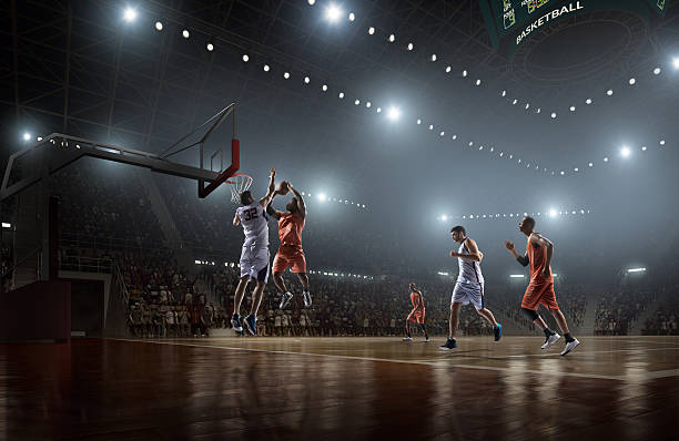 Basketball game Low angle view of a professional basketball game. A player is in mid air holding ball about to score a slam dunk, but the player from the opposite team is ready to block him.  A  game is in a indoor floodlit basketball arena. All players are wearing generic unbranded basketball uniform. basketball stock pictures, royalty-free photos & images