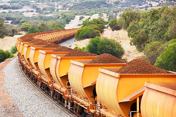 Heavy train loaded with brown hematite iron ore in orange freight wagons (cars) passes through rolling hills.  Horizontal, telephoto, ID removed, copy space.